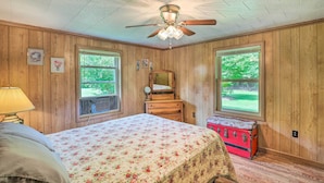 Een slaapkamer met houten wanden, een bed met bloemenbeddengoed, een plafondventilator, een dressoir, een spiegel en een koffer.