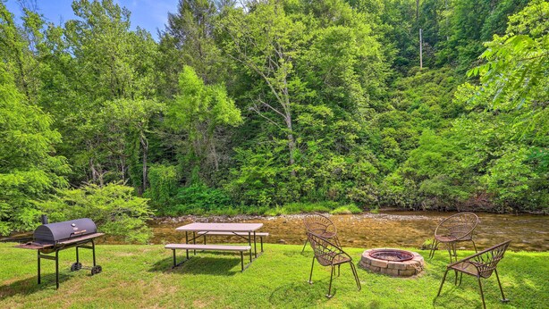Een terras buiten met een barbecue, picknicktafel, stoelen en een vuurkuil, omringd door weelderige begroeiing en een beek.