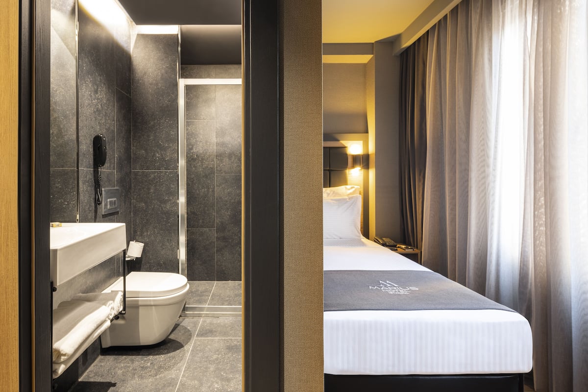 A modern hotel bathroom with a toilet, sink, and shower. The room features a bed with a headboard and a bedside table.