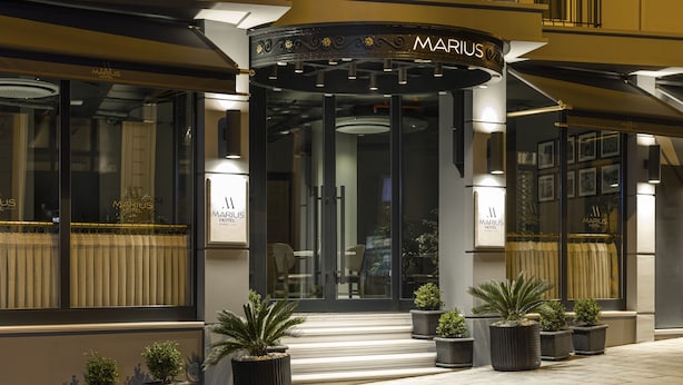 El exterior del Hotel Marius, con entrada de vidrio, plantas en macetas y fachada bien iluminada.
