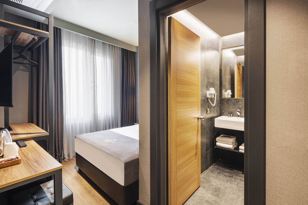 A hotel room with a bed, a desk with a chair, a wardrobe, a bathroom with a sink and mirror, and a shower.