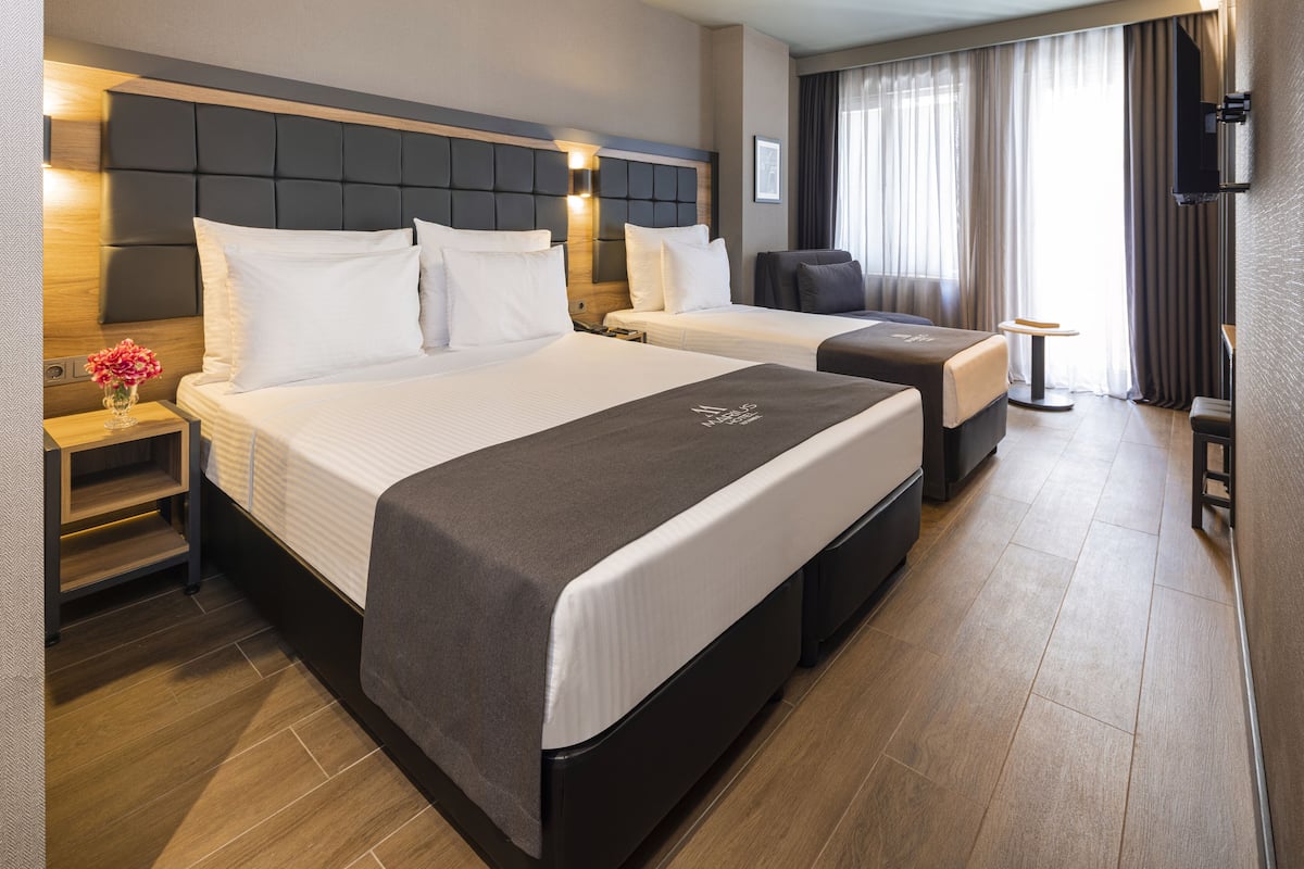 A hotel room with two beds, a nightstand with a vase of flowers, a chair, a small table, and a TV mounted on the wall.