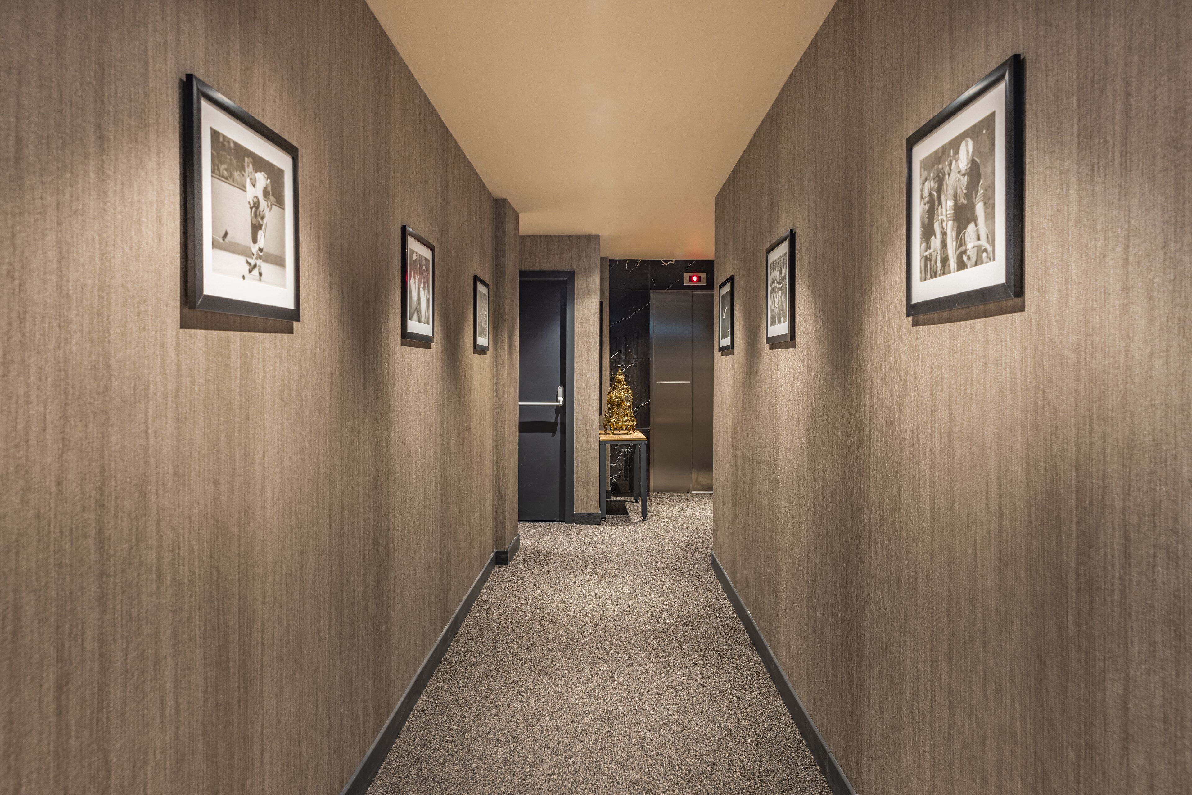 A hallway with framed pictures on the walls, a carpeted floor, and an elevator in the background.