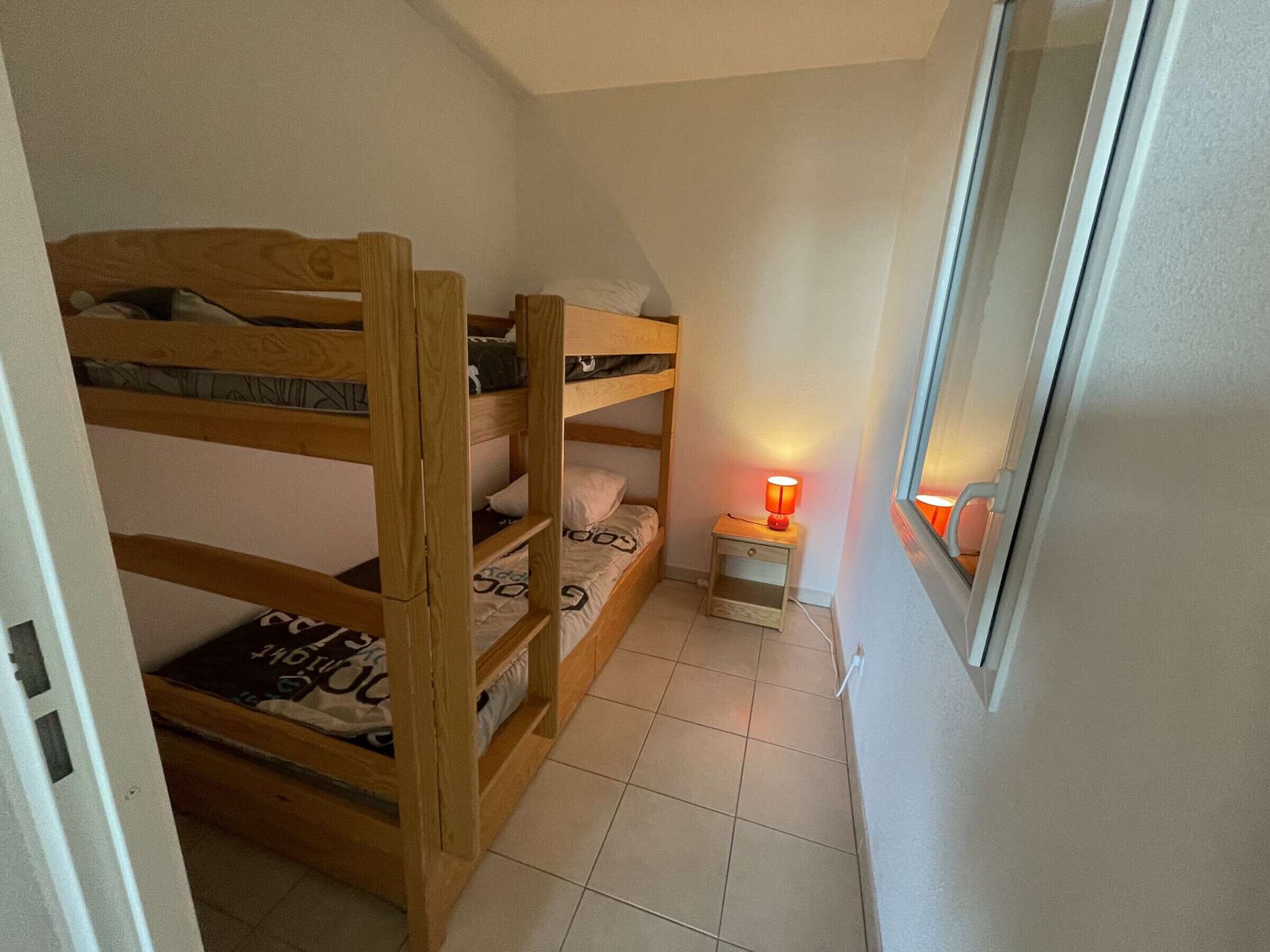 Habitació petita i estreta amb literes, una tauleta de nit amb una làmpada i una finestra.