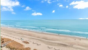 Ein Strand mit blauem Himmel, ruhigem Meer und einem sandigen Ufer, an dem einige Personen sind.
