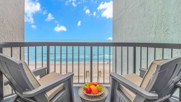 Ein Balkon mit zwei Stühlen, einem Tisch mit Früchten und Blick auf Strand und Meer.