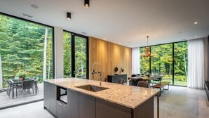 Una cocina moderna con una isla grande, paneles de madera y vistas a un bosque a través de amplios ventanales.