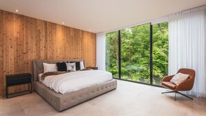 Un dormitorio con una cama grande, una mesita de noche, una silla y vistas a un bosque a través de amplios ventanales.
