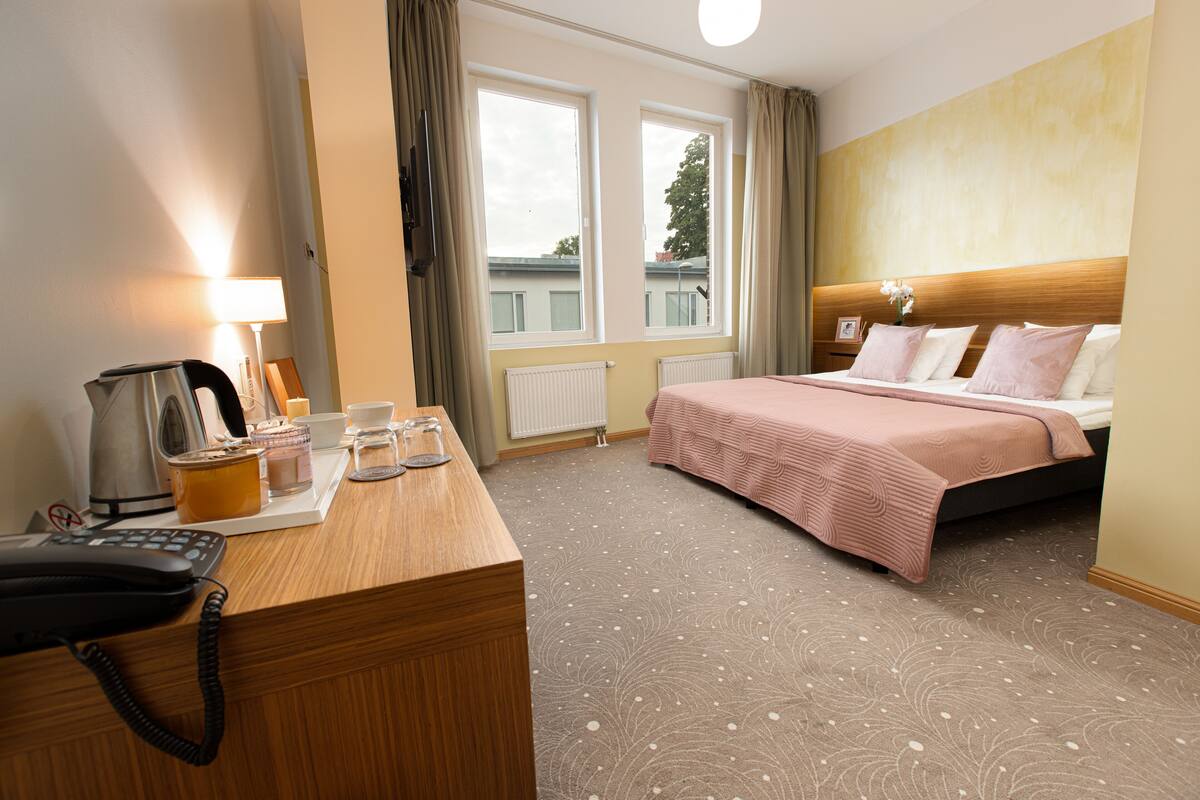 A hotel room with a wooden desk, a bed with a pink bedspread, a window with curtains, and a radiator.