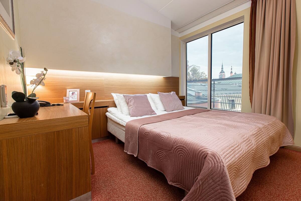 A hotel room with a bed, a desk, a chair, a large window with a view of buildings, and a potted orchid on the desk.