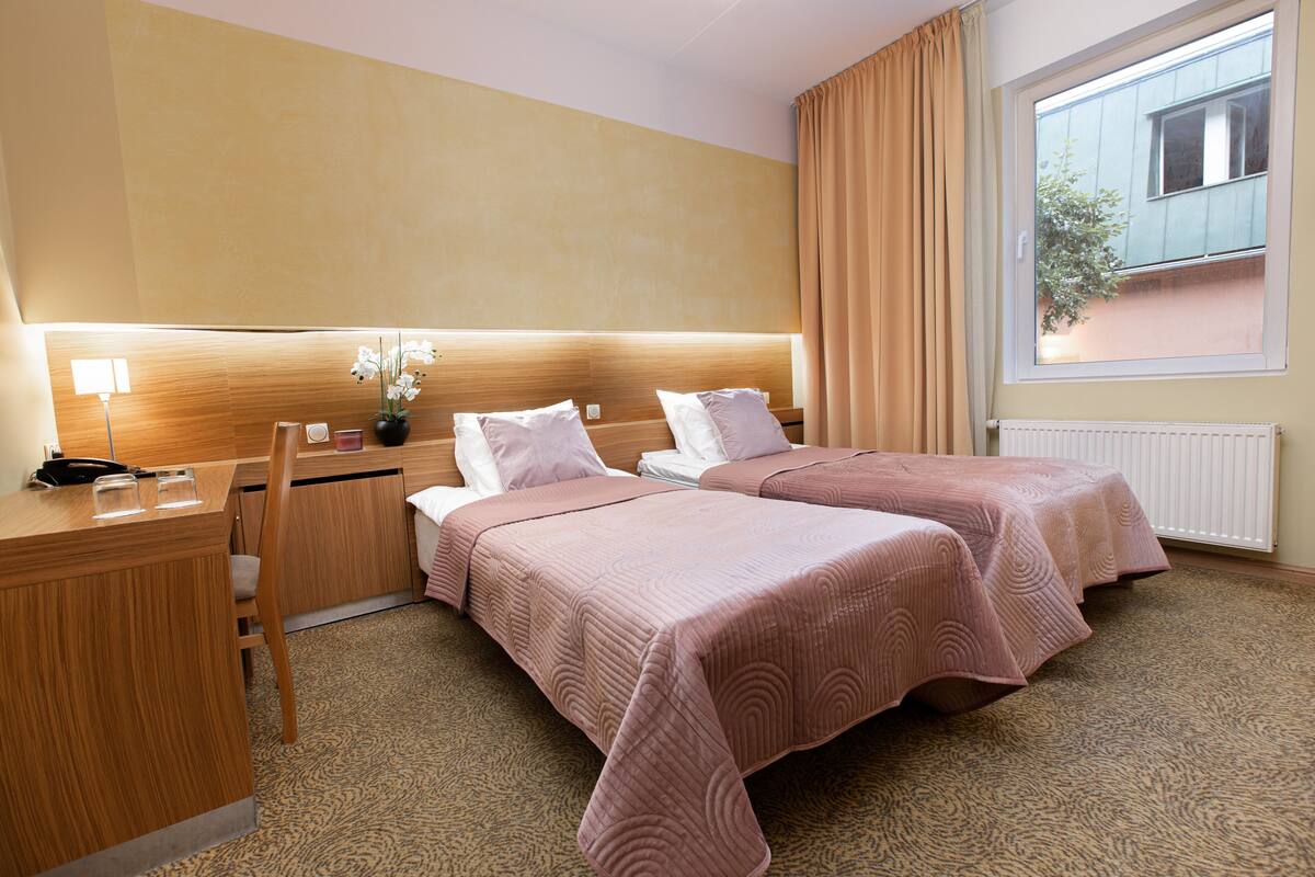 A hotel room with two beds, a desk, a chair, a lamp, and a window with curtains.