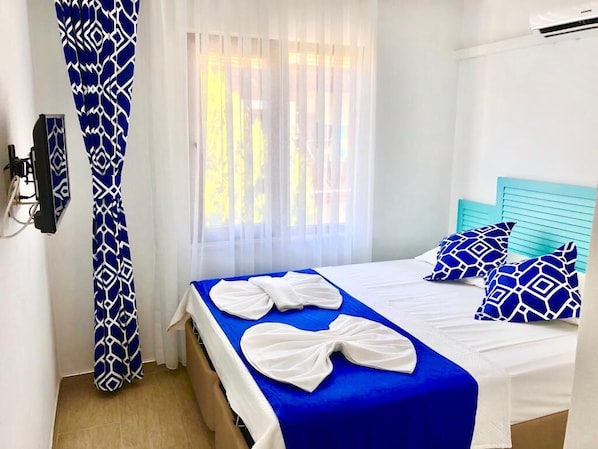 A hotel room with a bed, a blue and white patterned curtain, and a window with sheer curtains.