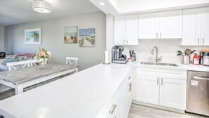 Een moderne keuken met witte kasten, een roestvrijstalen spoelbak en een ingebouwde oven. Er is een zithoek met een witte tafel en stoelen, en kunstwerken aan de muren.