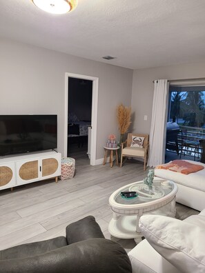 Ein modernes Wohnzimmer mit Flachbildfernseher, weißen Sofas, einem Couchtisch und Blick ins Freie.