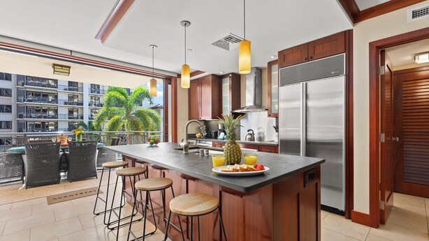 Una cocina moderna con una isla central, taburetes de bar, electrodomésticos de acero inoxidable y vistas a un edificio residencial.