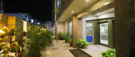 A well-lit outdoor corridor with potted plants, a doormat, and a door leading to a room with a view.