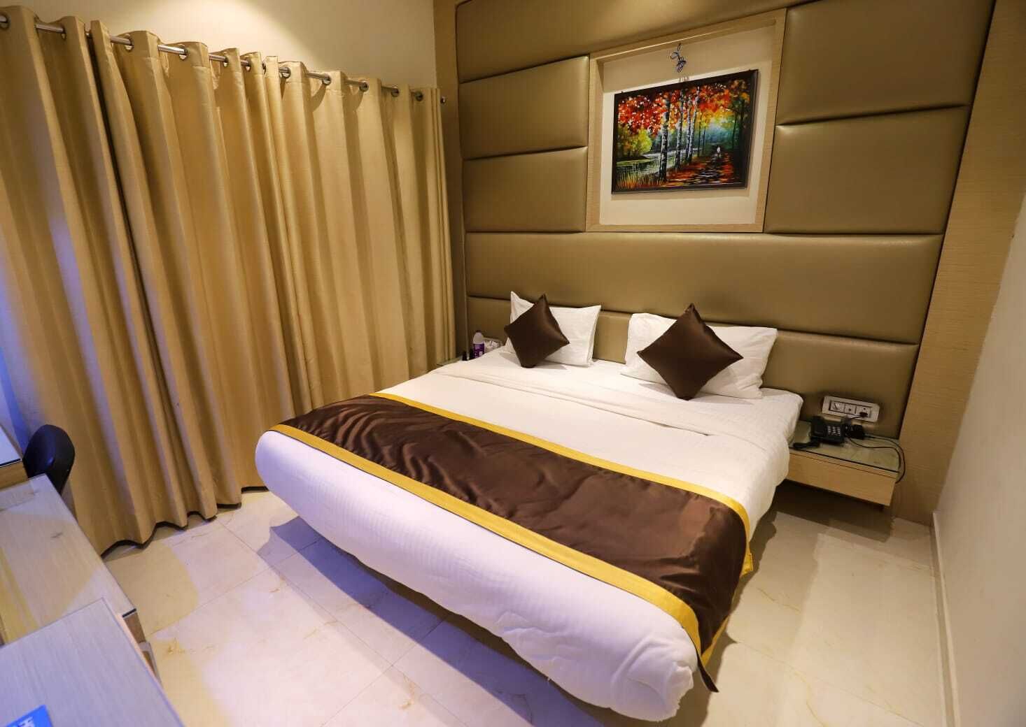 A hotel room with a large bed, beige curtains, a painting on the wall, and a bedside table with a phone and charger.