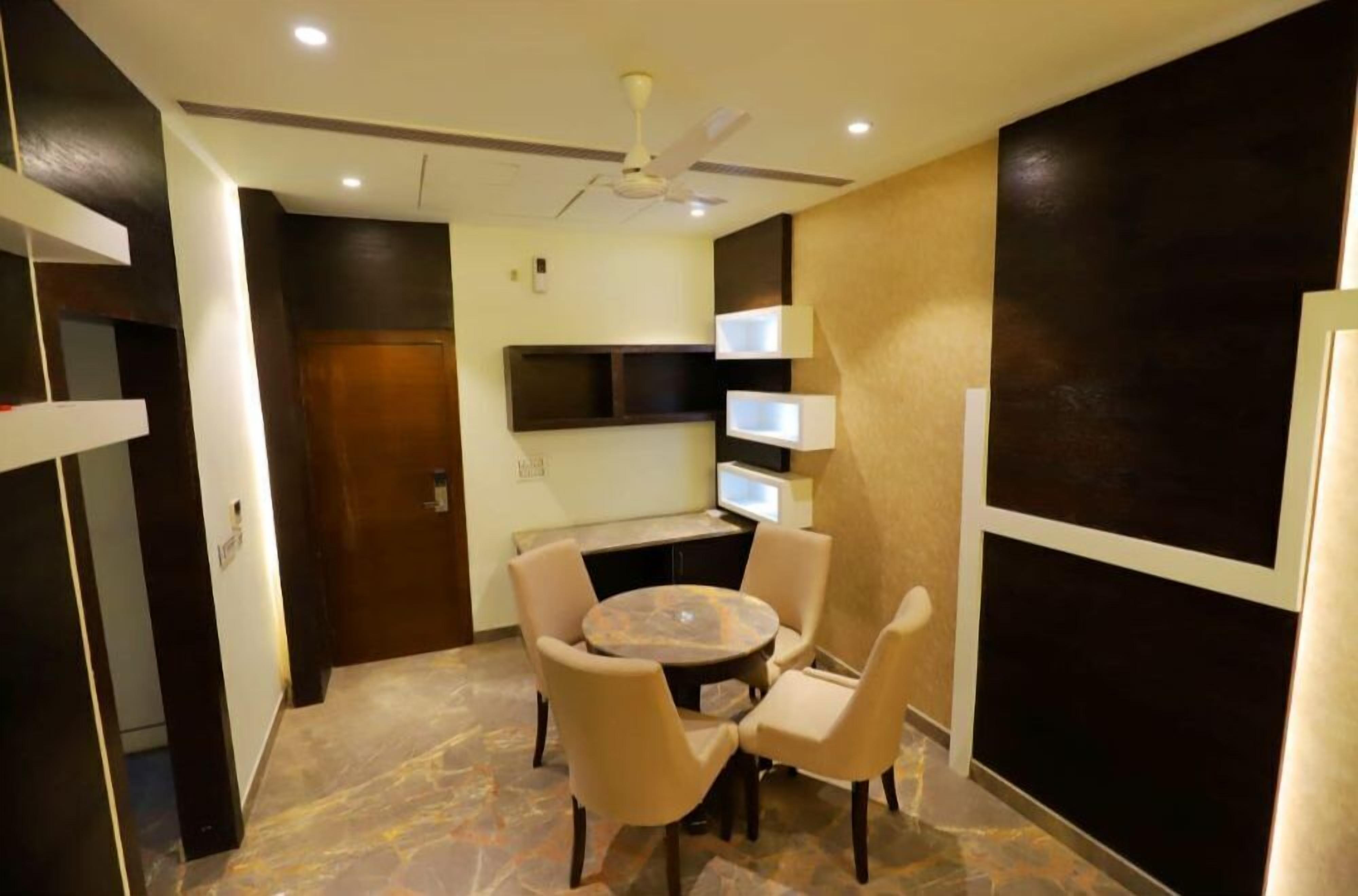A hotel room with a round table and four chairs, a ceiling fan, a TV mounted on the wall, and a wooden door.