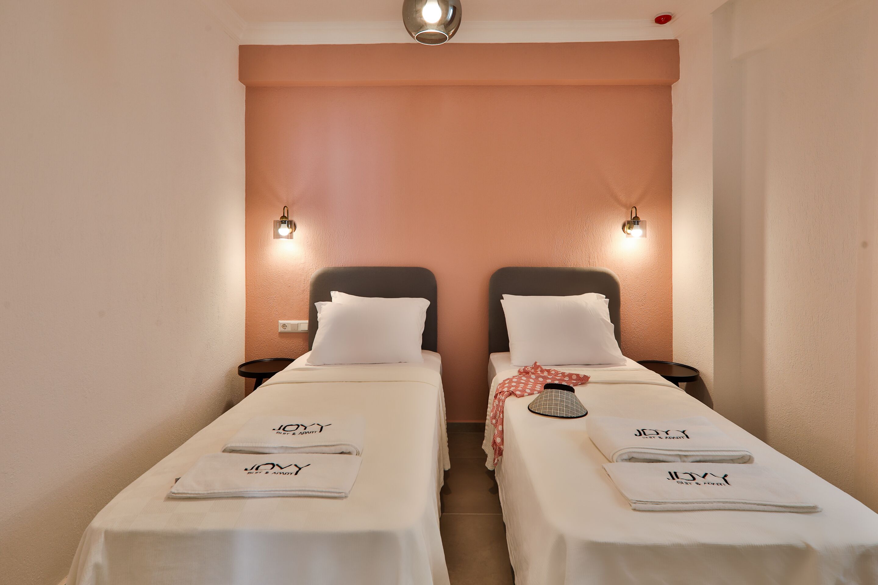 A hotel room with two single beds, each with a white bedspread and matching pillows. There are two bedside tables, one on each bed, with a lamp on each. The walls are painted in a warm peach color, and there is a ceiling light fixture above the beds.