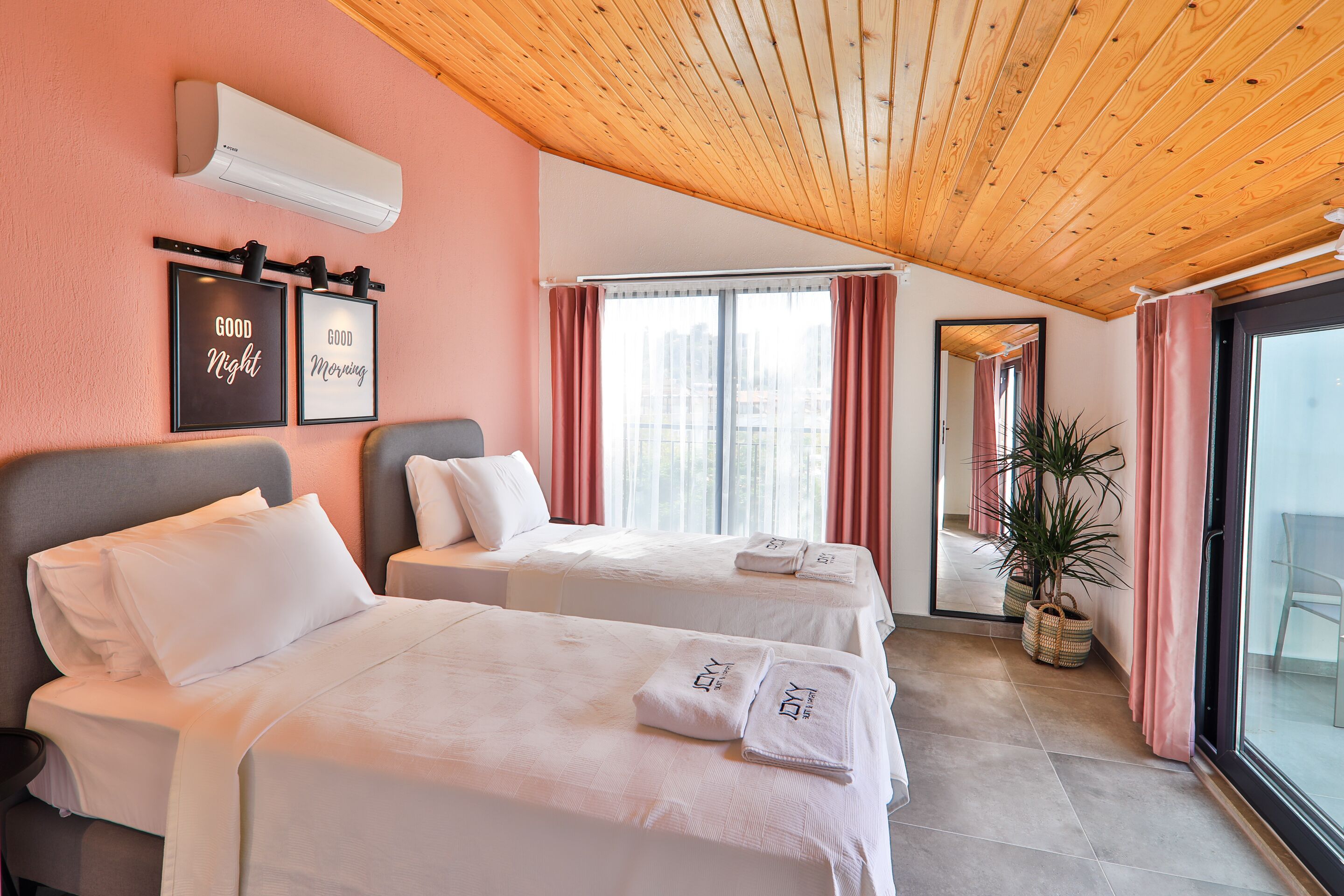 A hotel room with two beds, a large window with curtains, a ceiling fan, and a potted plant.