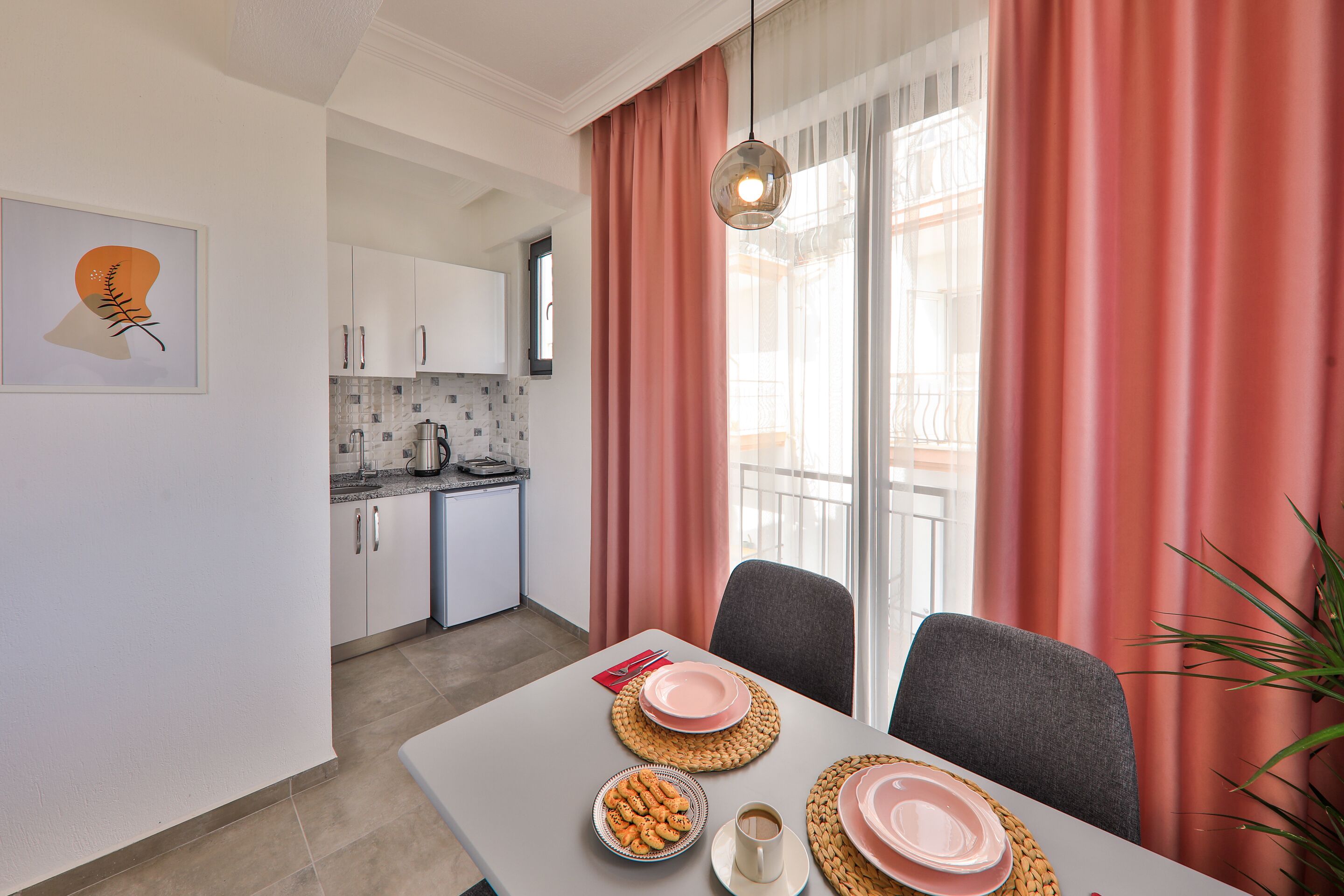 Un coin repas avec une table dressée pour deux personnes, une cuisine en arrière-plan et une grande fenêtre avec des rideaux.