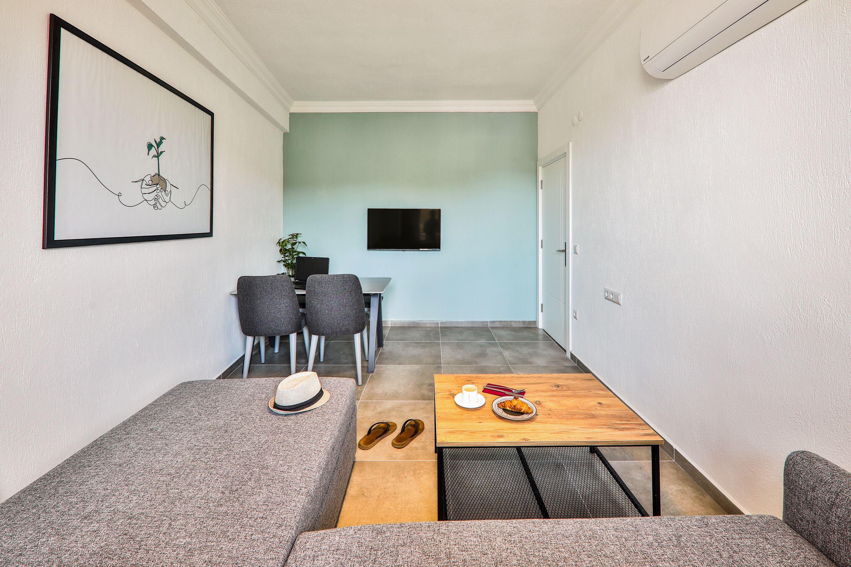 Un espacio habitable compacto con un sofá, una mesita con un plato de comida, un par de zapatillas, un cuadro enmarcado y un televisor montado en la pared.