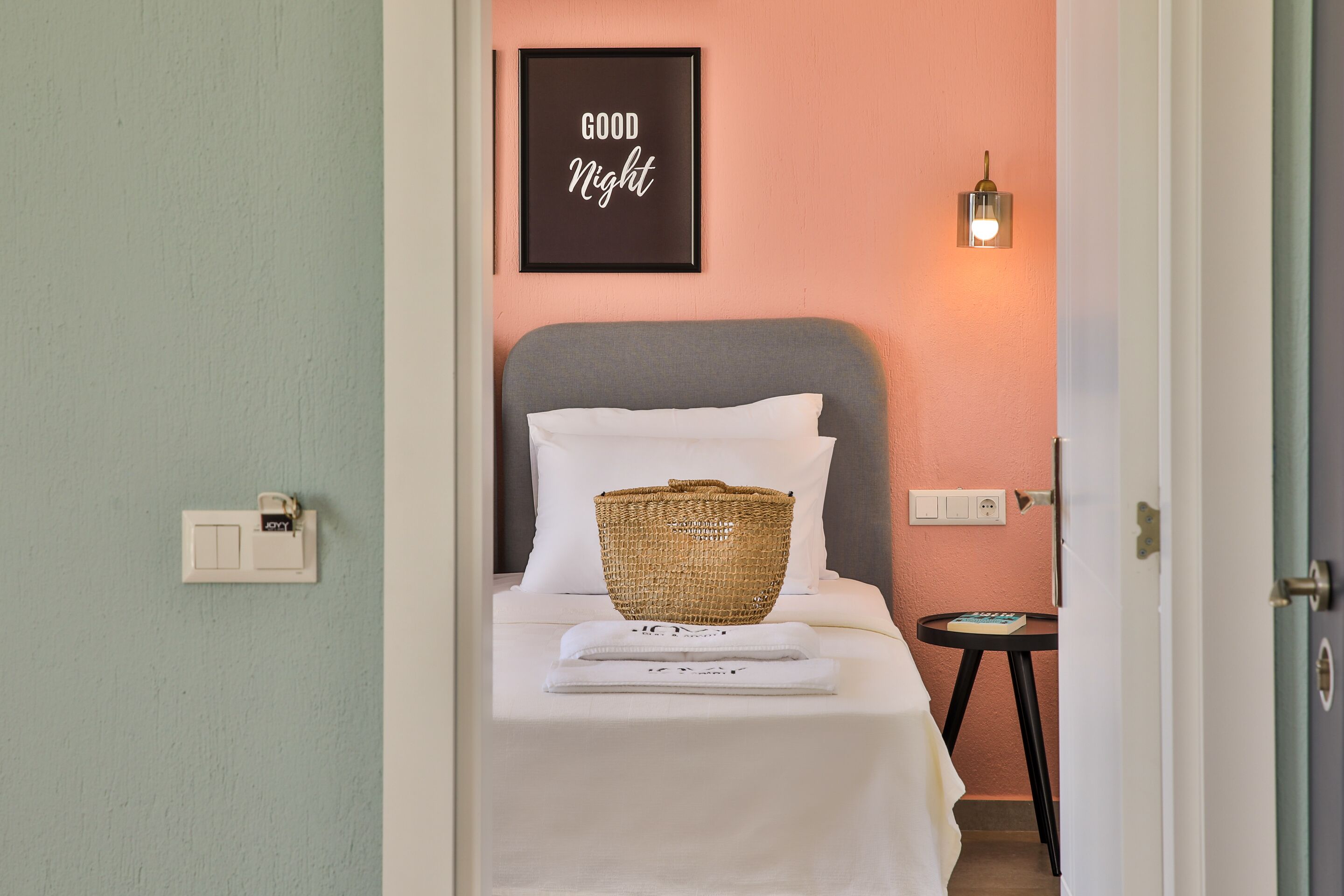 Une chambre d’hôtel avec un lit, un panier, un panneau encadré portant l’inscription « BONNE NUIT » et une lampe fixée au mur.