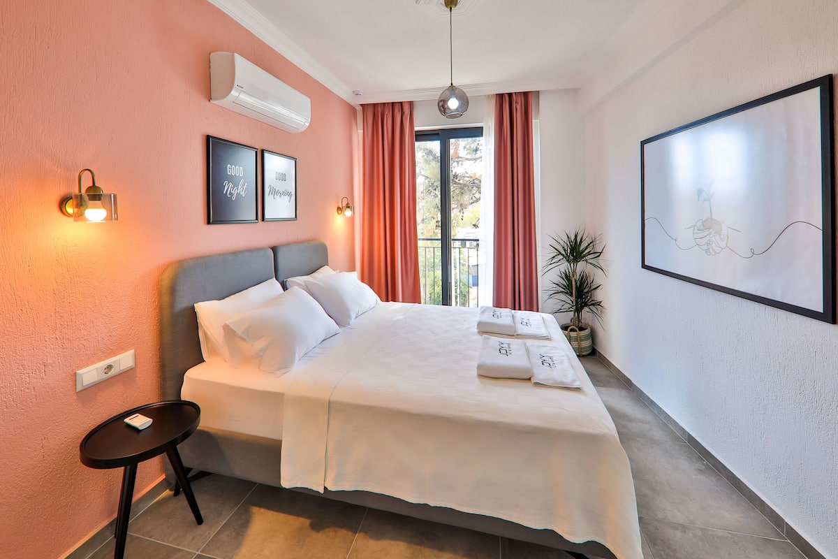 Uma cama bem arrumada com roupa de cama e toalhas brancas, uma pequena mesa de cabeceira, um ar-condicionado fixado na parede e uma obra de arte emoldurada acima da cama.