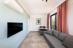 Una sala de estar con una televisión de pantalla plana, un sofá gris, una mesa de comedor con sillas y una ventana con cortinas rojas.