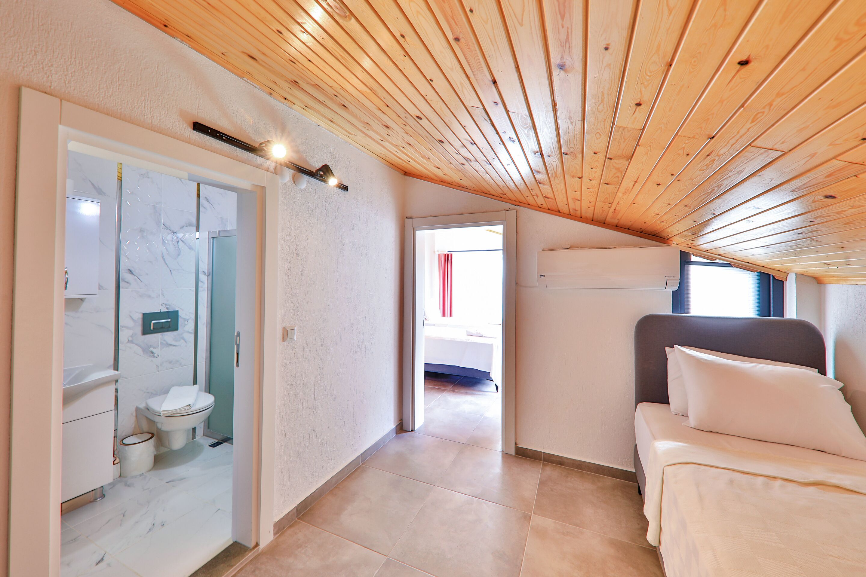 A bedroom with a wooden ceiling, a bed with white bedding, and a bathroom with a marble floor and white fixtures.