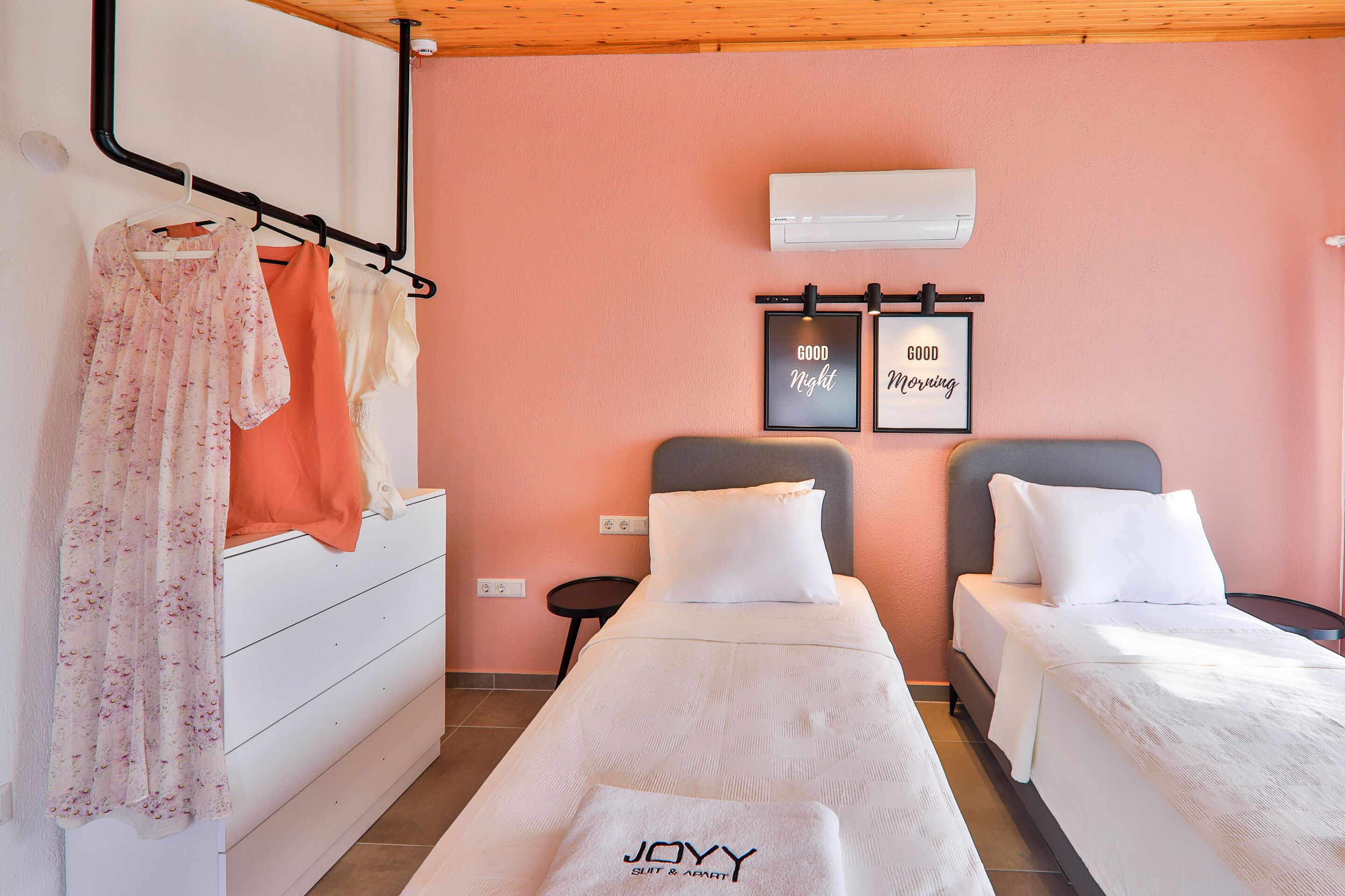 A bedroom with two beds, a white dresser, a hanging rack with clothes, and two framed signs saying "Good Night" and "Good Morning".