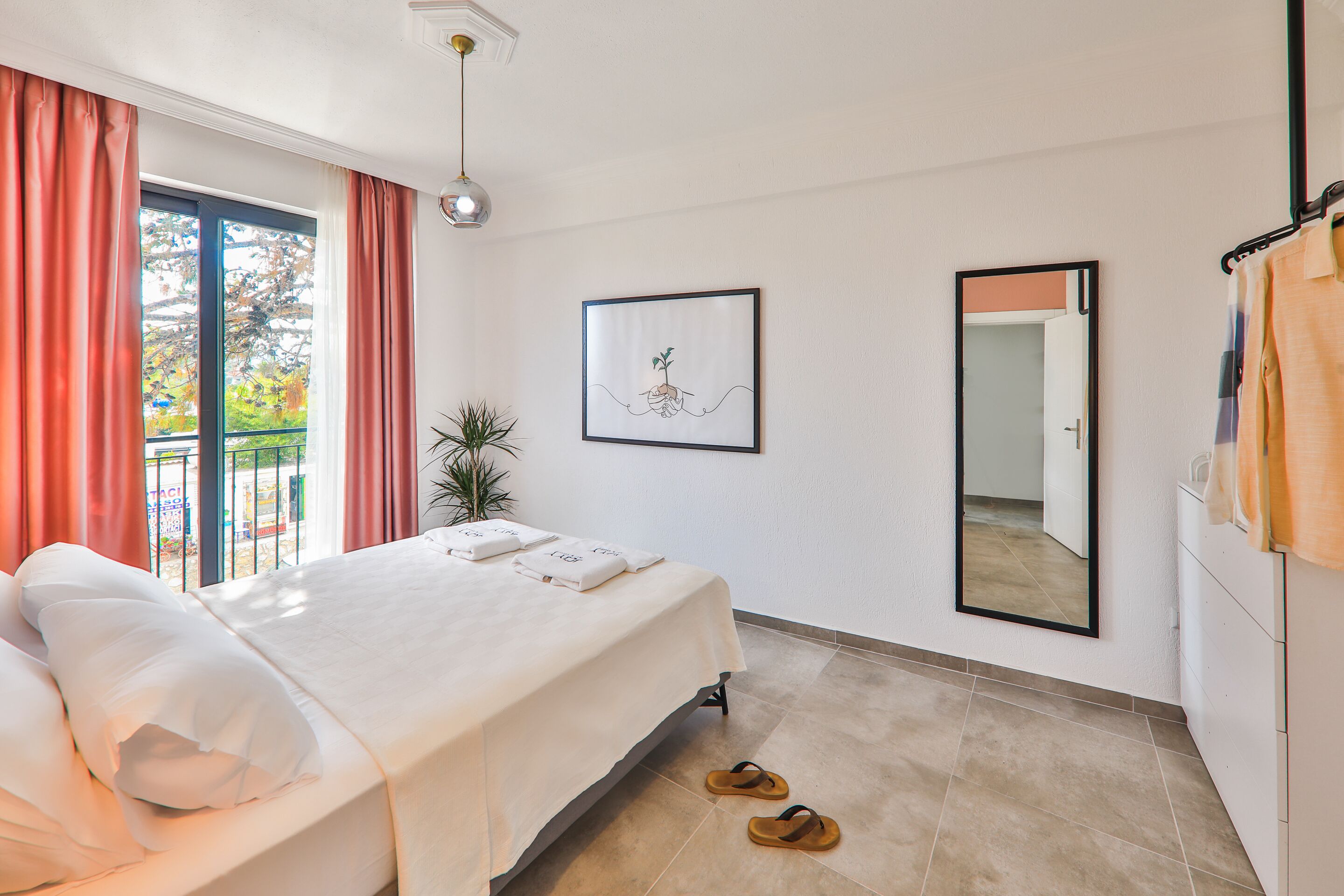 Une chambre d’hôtel avec un lit, une fenêtre avec des rideaux, un tableau encadré au mur, un miroir et une paire de chaussons sur le sol.