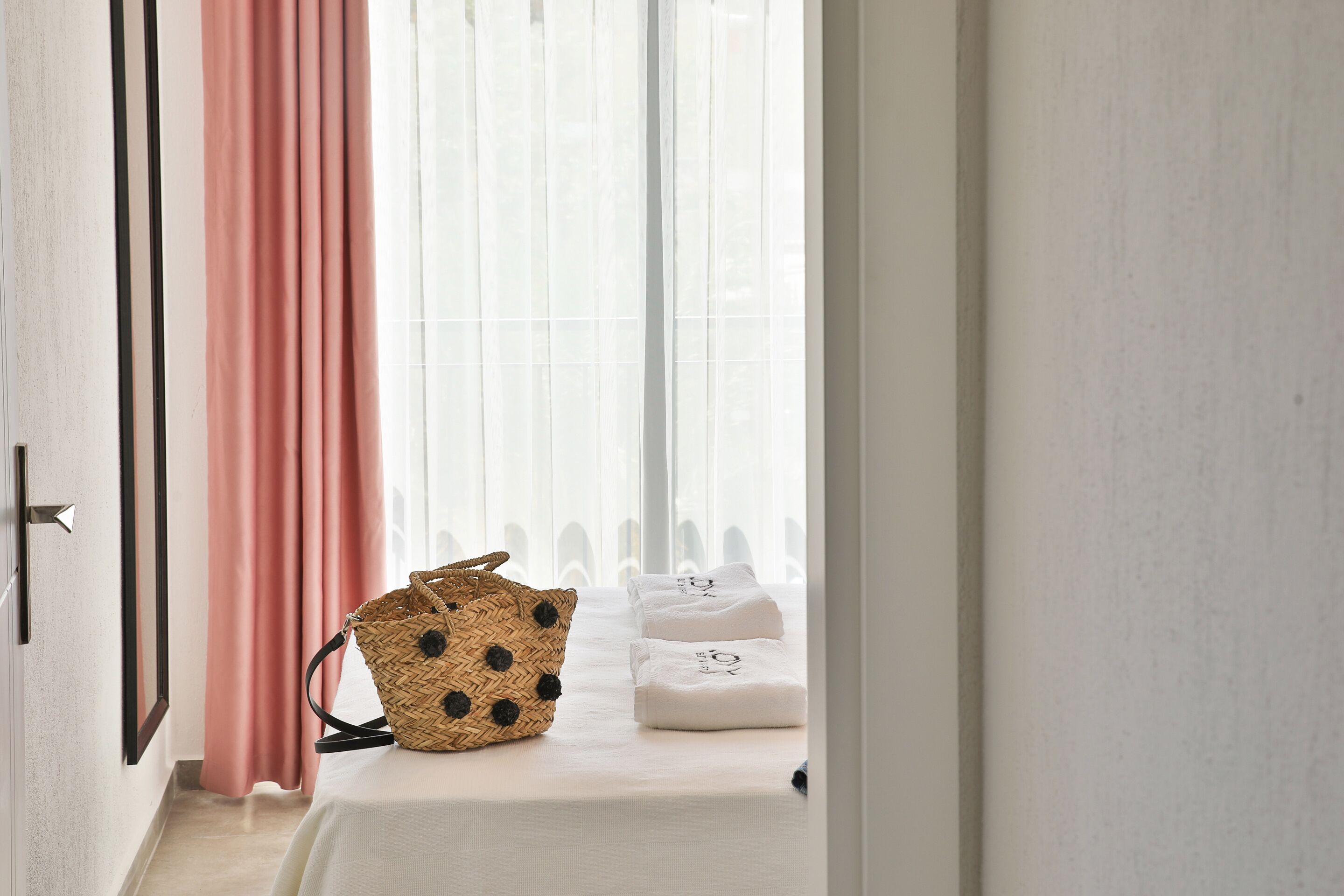 A hotel room with a bed, a woven bag, and a window with sheer curtains.