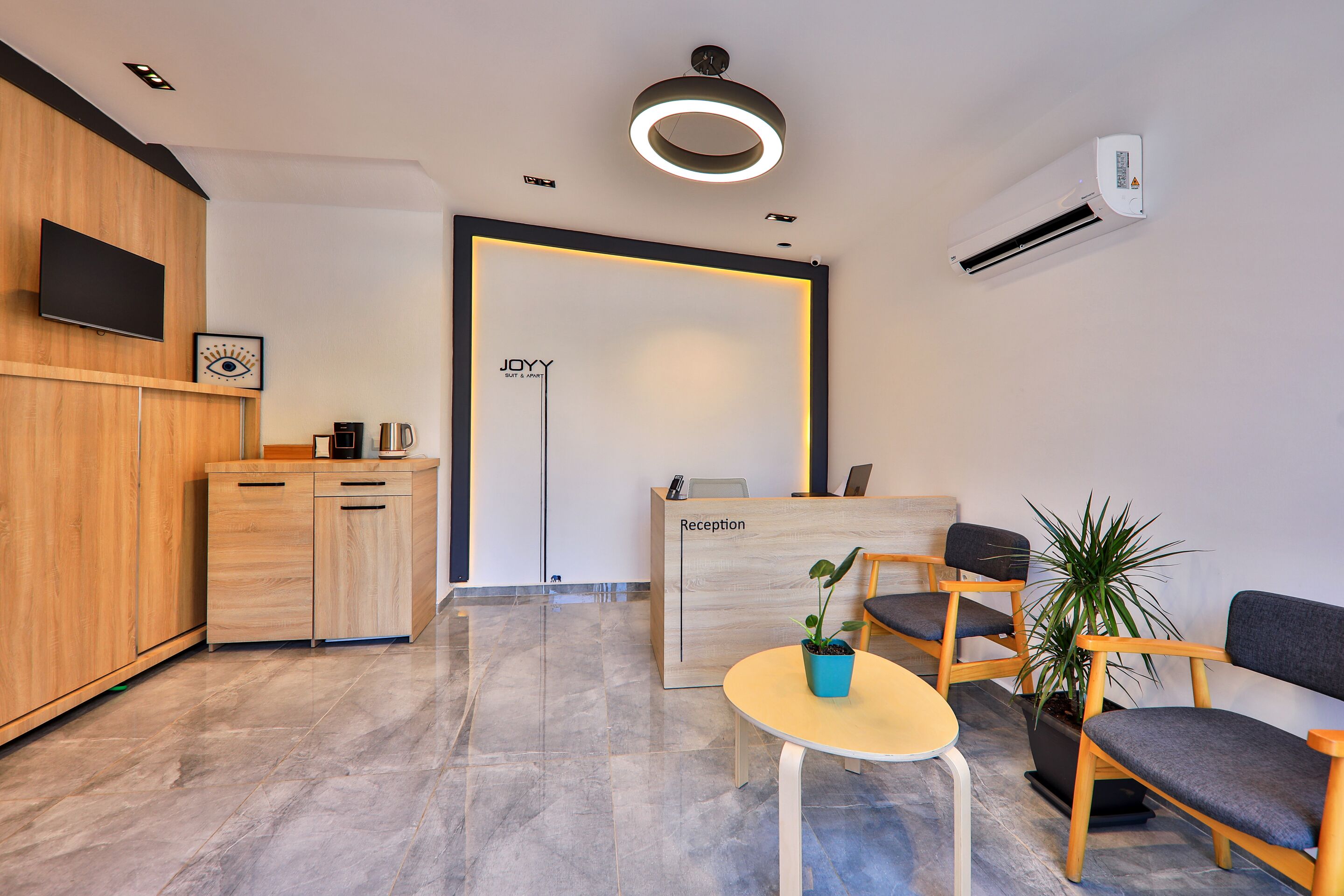 Un espace d’accueil avec un bureau en bois, des chaises, une petite table avec une plante et un téléviseur fixé au mur.