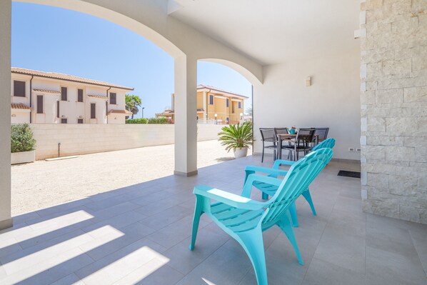 A covered outdoor seating area with turquoise chairs, a table, and a potted plant.