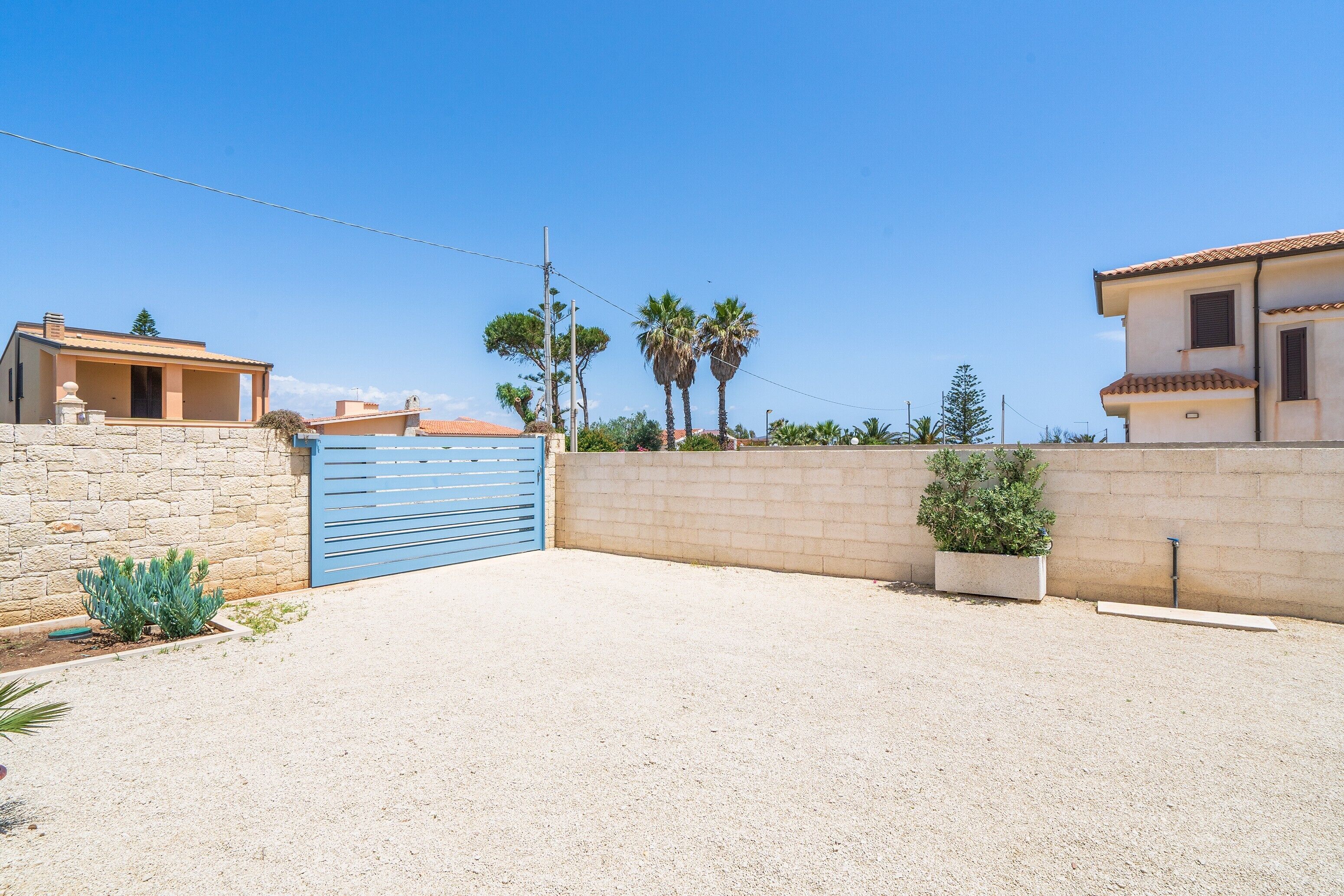 Uma área residencial com entrada de automóveis em cascalho, um portão azul e uma parede de pedra.