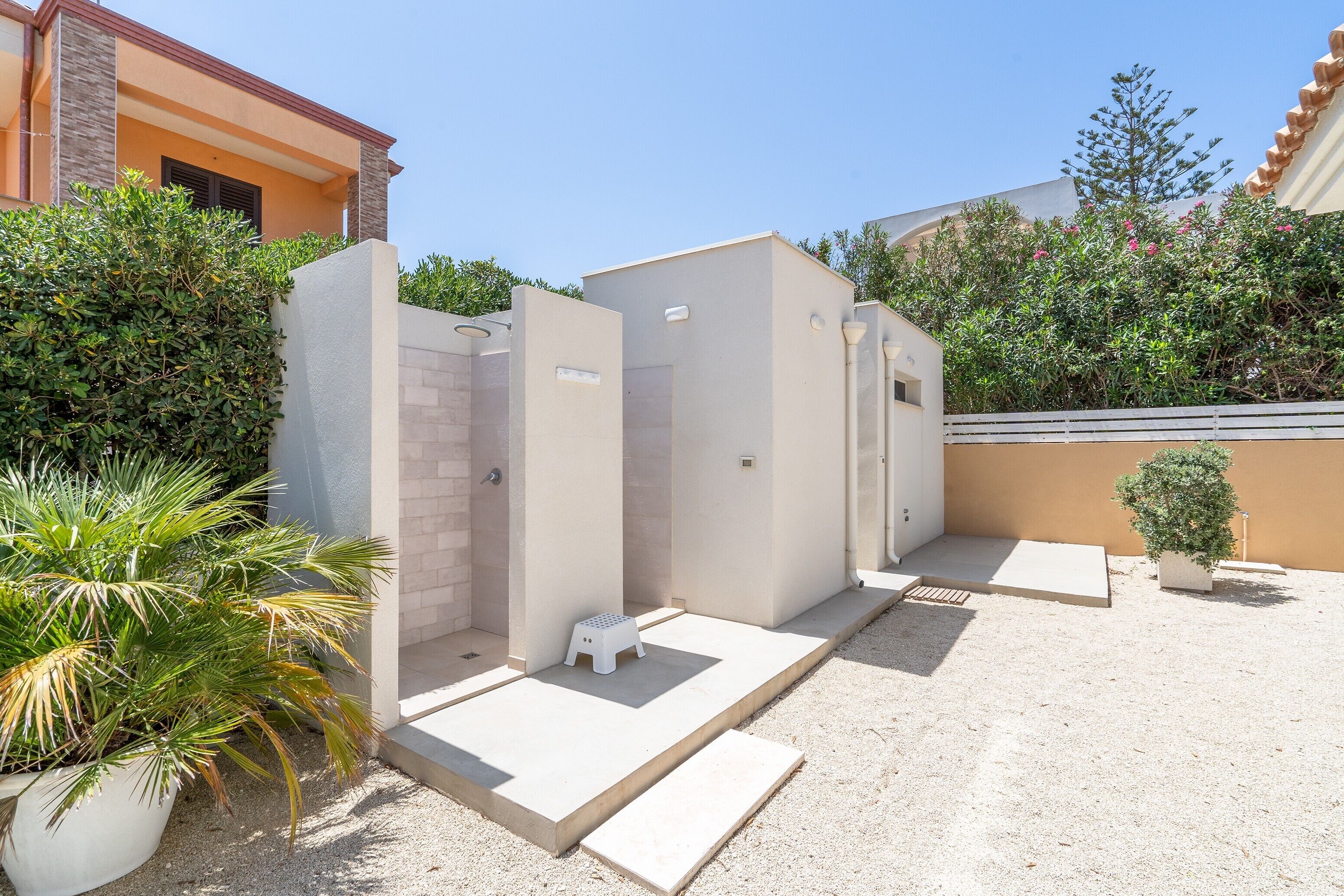 A modern outdoor restroom area with a small stool, a planter with a palm plant, and a gravel ground.