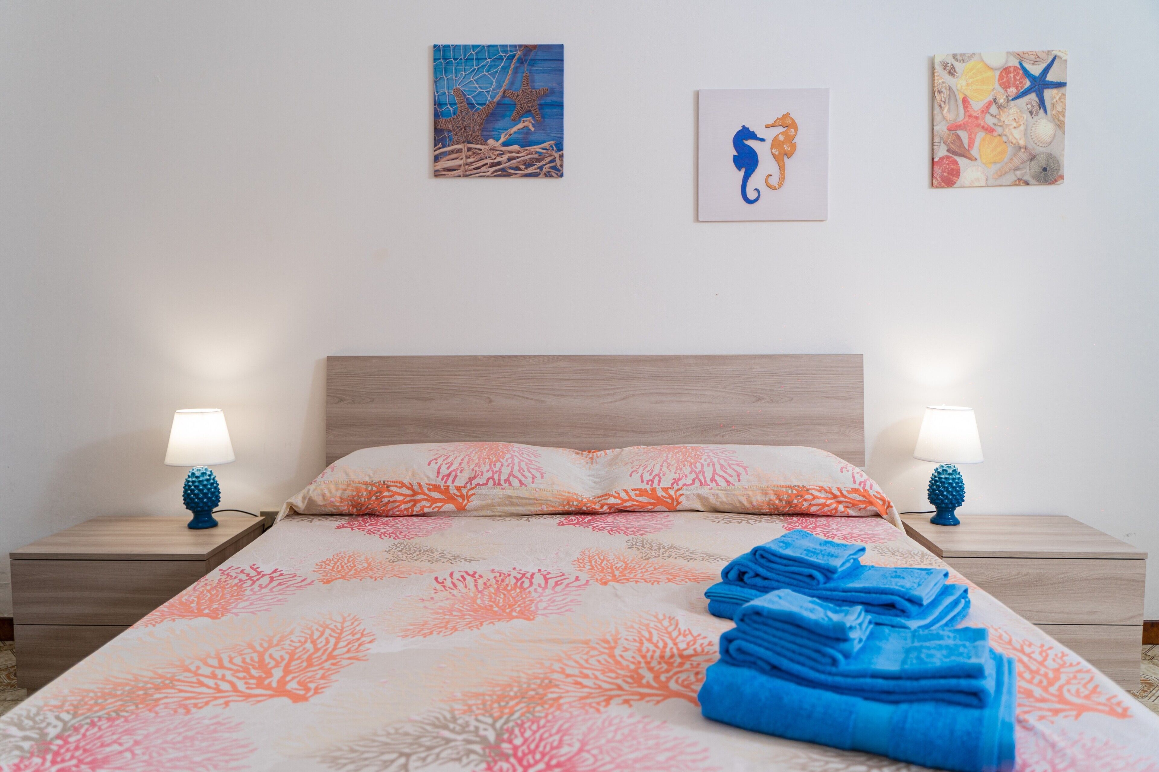 A neatly made bed with a coral-patterned bedspread, two bedside lamps, and three wall-mounted artworks depicting marine life.