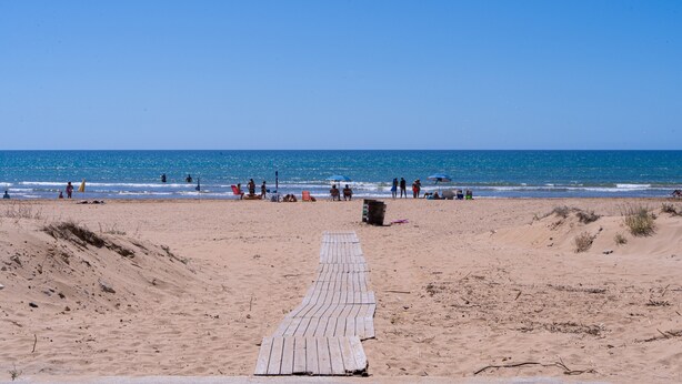 Una playa con un pasillo de madera que lleva al agua, personas a lo lejos y un cielo despejado.