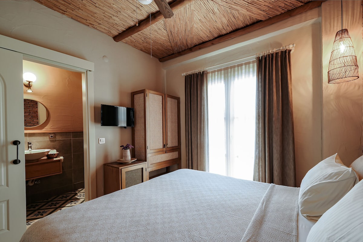 Habitación de hotel con cama, televisor, armario de madera, baño con lavamanos y ventana con cortinas.