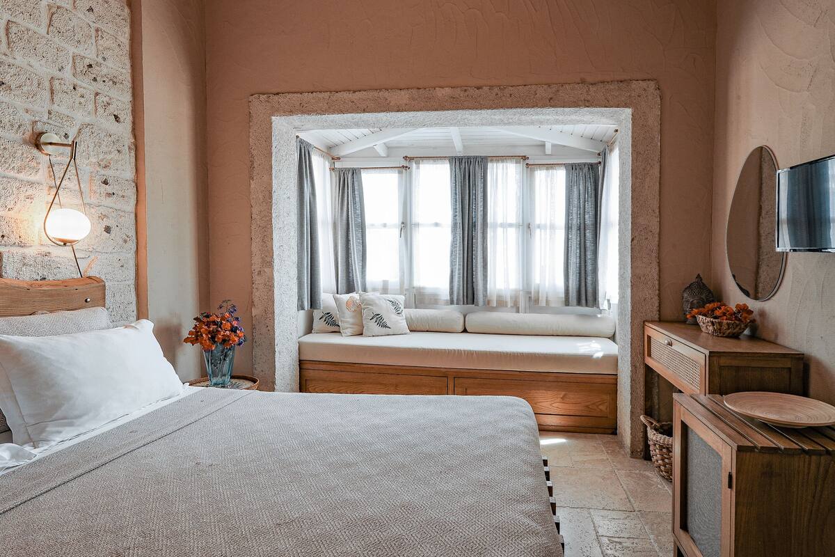 Un dormitorio con una cama grande, un rincón con asiento junto a la ventana, un aparador de madera y un televisor de pared.