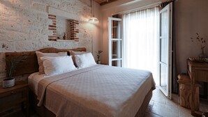 A bedroom with a brick wall, a wooden headboard, a large bed with white bedding, a bedside table with a plant, and a window with curtains. - Lika Alacati (Cesme)
