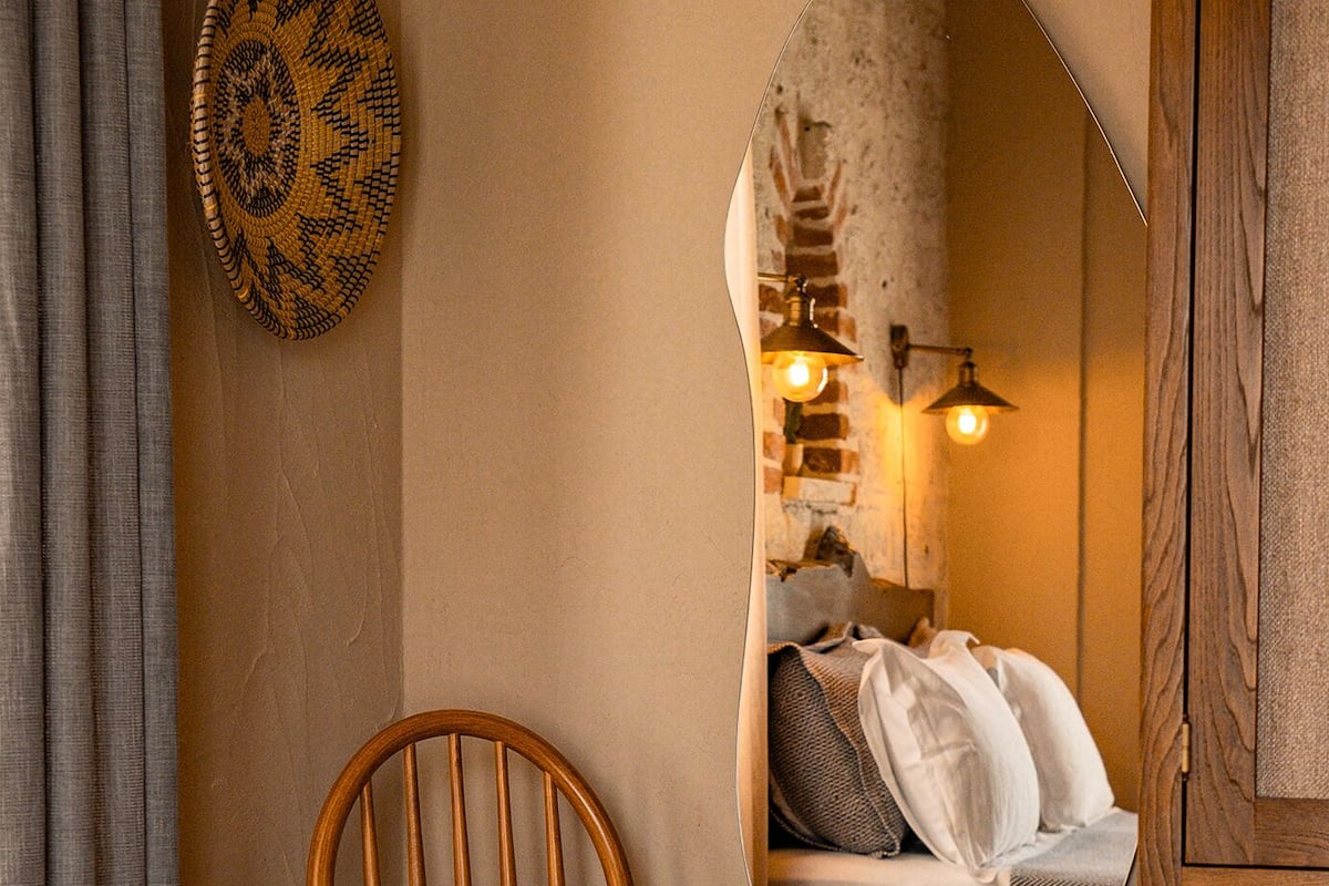 Un dormitorio con una silla de madera, una cama con ropa de cama blanca, un espejo que refleja la habitación y un aplique de pared.