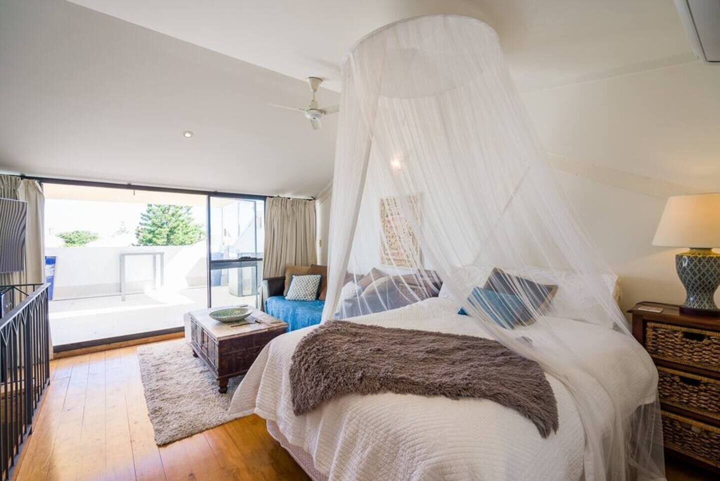 A bedroom with a canopy bed, a view to the outside, a small table, and a lamp.