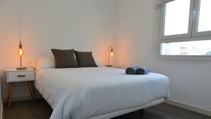 A bedroom with a bed, two bedside tables with lamps, a window, and a view of buildings. - Holiday Home Marina with Mountain View, Terrace & Wi-Fi (Las Palmas de Gran Canaria)