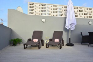 Duas cadeiras de descanso com toalhas, uma mesinha com dois copos, um vaso de planta e um guarda-sol branco em um terraço.