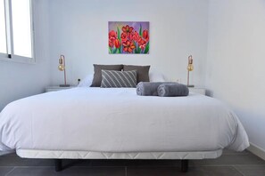Uma cama grande com roupa de cama branca, duas luminárias de cabeceira, um quadro de flores e uma janela à esquerda.
