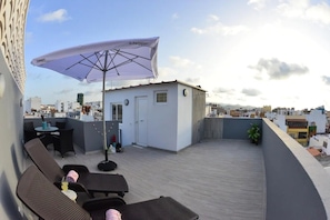 En tagterrasse med en lille hvid bygning, et bord med stole, en stor parasol og en potteplante.