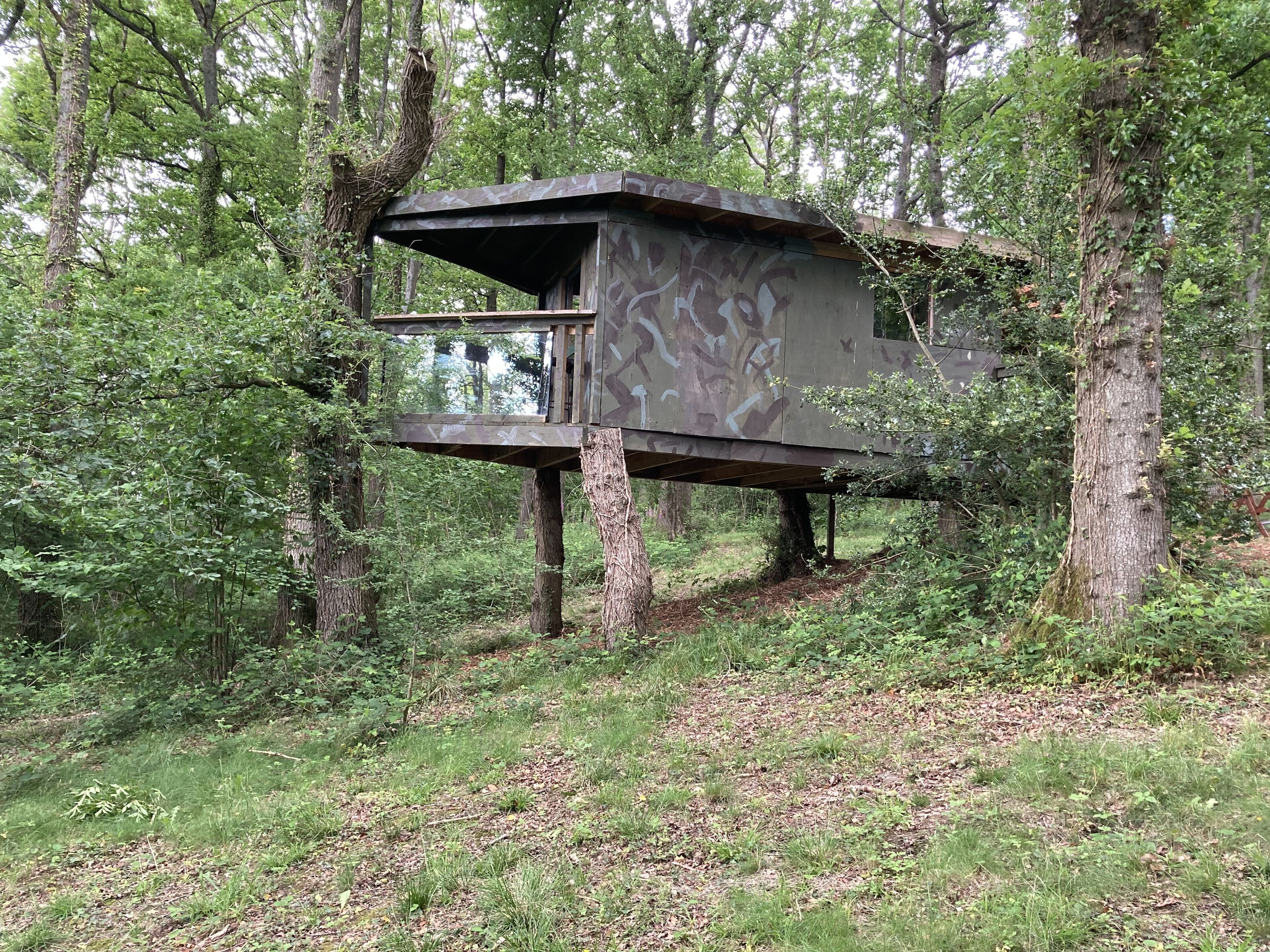 A treehouse with a camouflage exterior, built on a single tree trunk, surrounded by dense forest.
