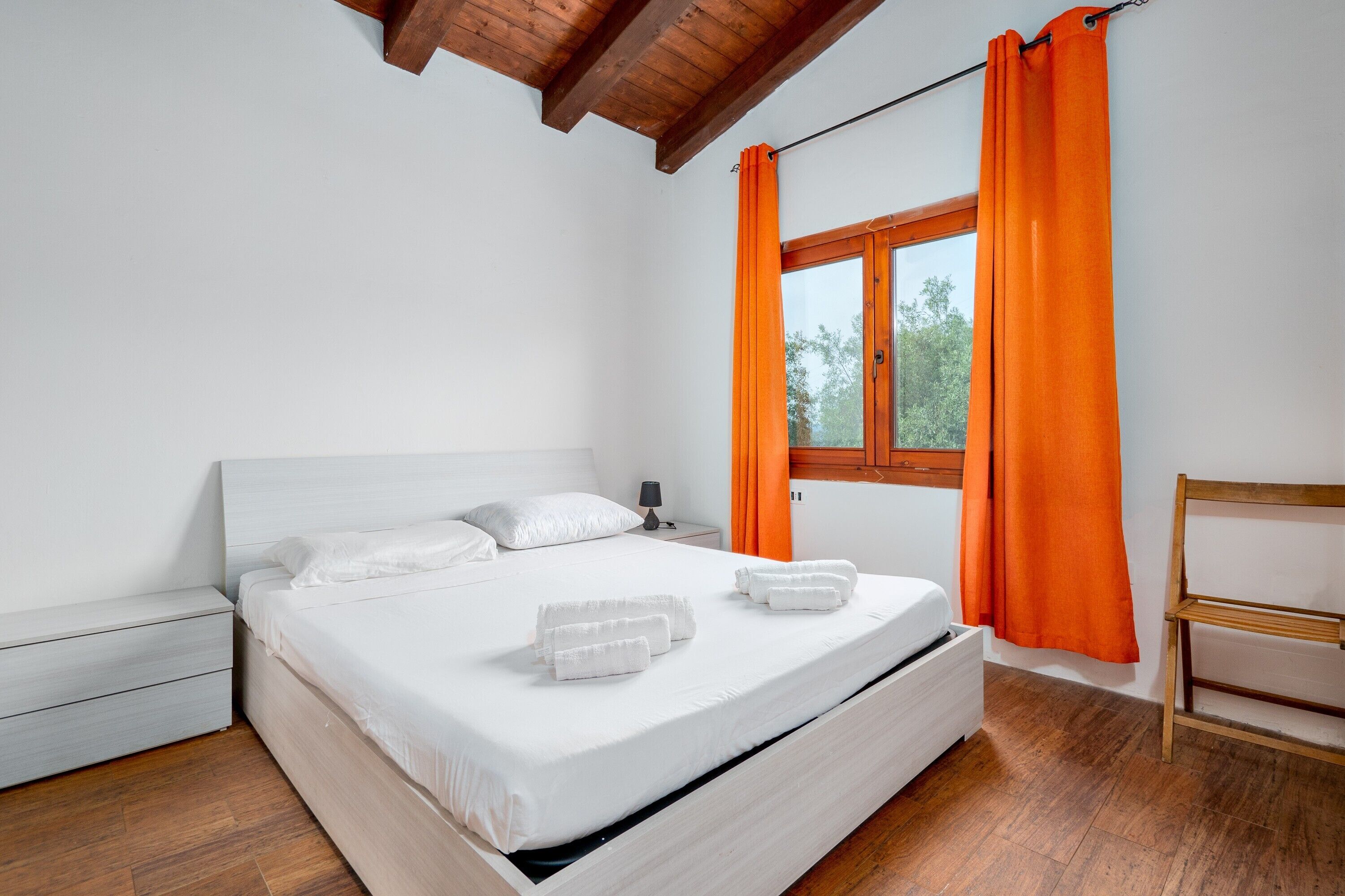 A bedroom with a bed, white bedding, a wooden nightstand, a wooden chair, and a window with orange curtains.